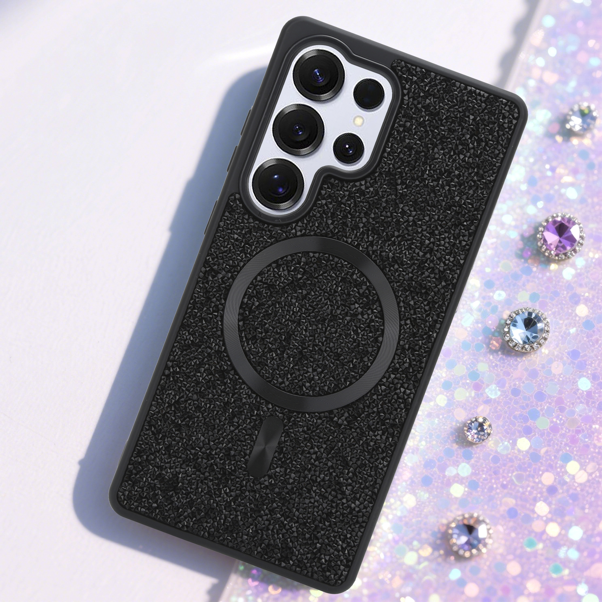 Schwarze Telefonhülle mit strukturierter Oberfläche, einem Magnetring und mehreren Kameralinsen. Glitzernder Hintergrund mit Edelsteinen.