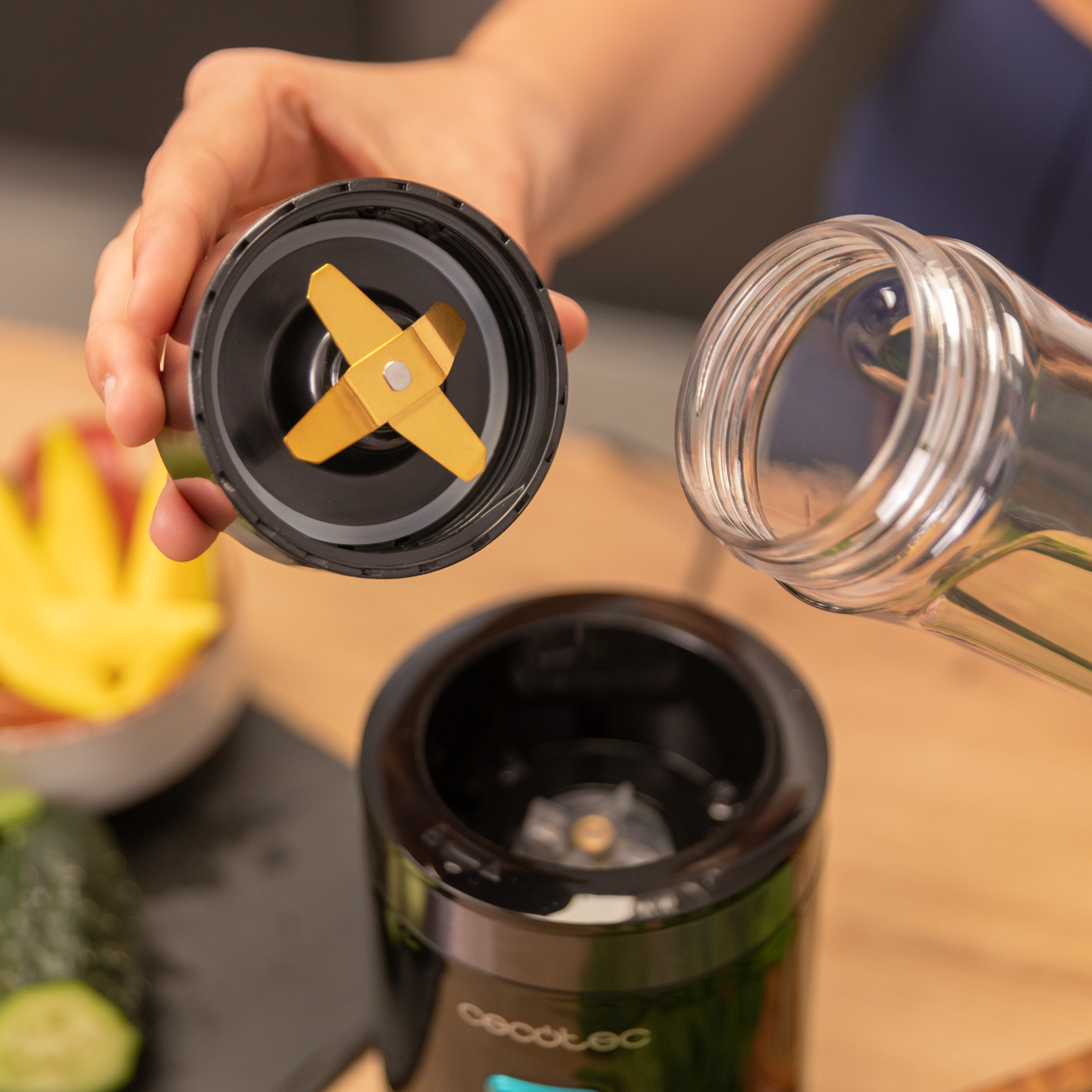 Een persoon houdt een blenderdeksel met messen boven de zwarte basis. Een doorzichtige fles wordt in de buurt gehouden. Fruit op de achtergrond.
