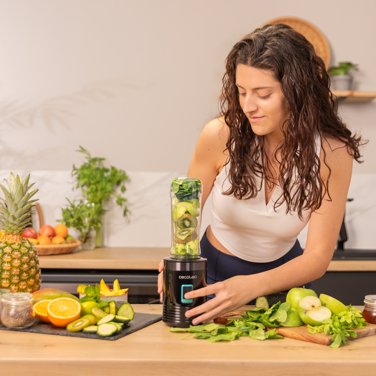 Vrouw maakt smoothie met blender, fruit en groenten op een houten aanrecht in de keuken. Ananas en mango zijn zichtbaar.
