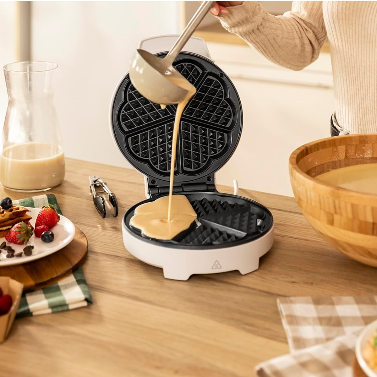 Una persona versa l'impasto in una macchina per waffle. Waffle, frutta e una ciotola di pastella su un tavolo di legno.
