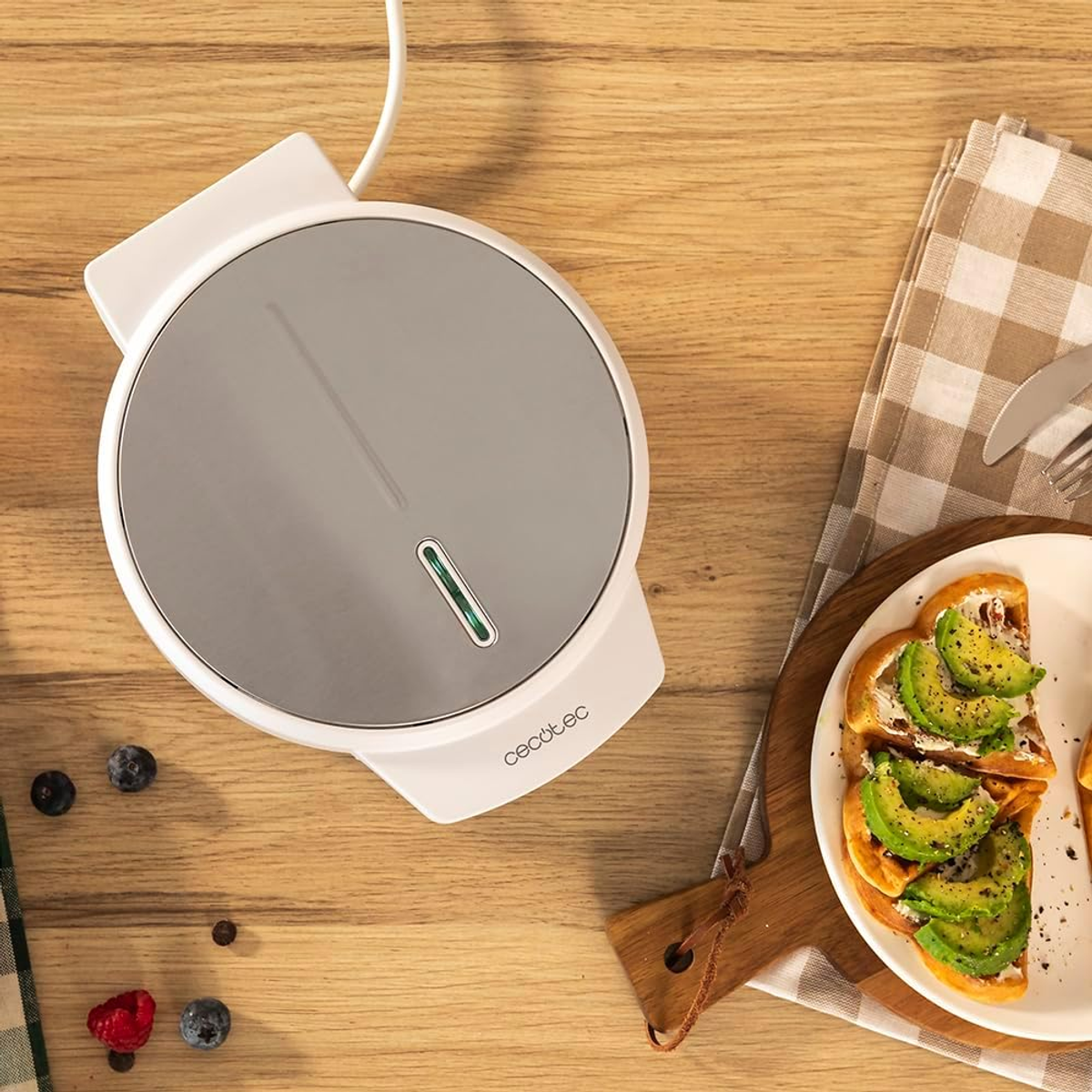 Una macchina per waffle bianca è su un tavolo di legno vicino a toast di avocado. Sono presenti anche bacche e un tovagliolo a scacchi.