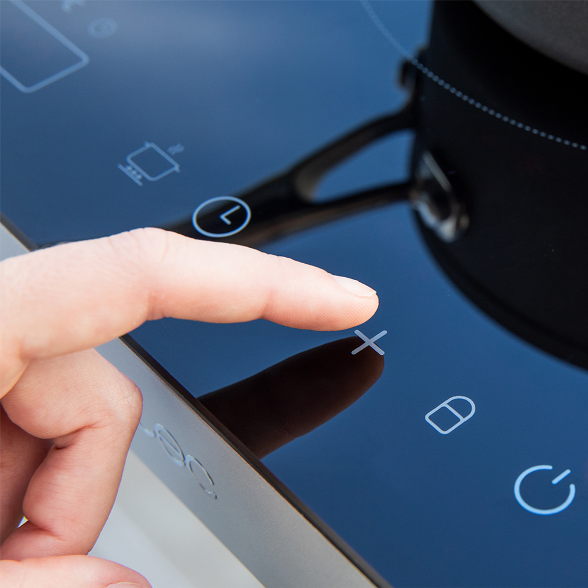 Palec naciska symbol „X” na czarnej płycie kuchennej. Garnek stoi na palniku. Widoczne są inne symbole.