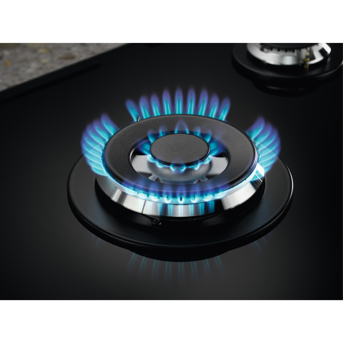 Nahaufnahme eines Gasherdbrenners mit blauen Flammen. Der Brenner ist schwarz mit einem silbernen Ring. Ein weiterer Brenner ist im Hintergrund sichtbar.