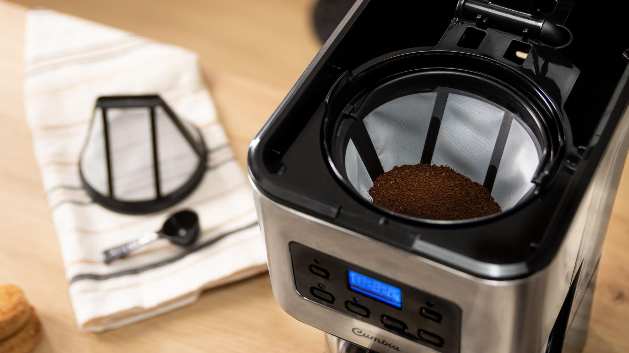 Ekspres do kawy z mieloną kawą w filtrze, obok ręcznika, filtra i miarki.