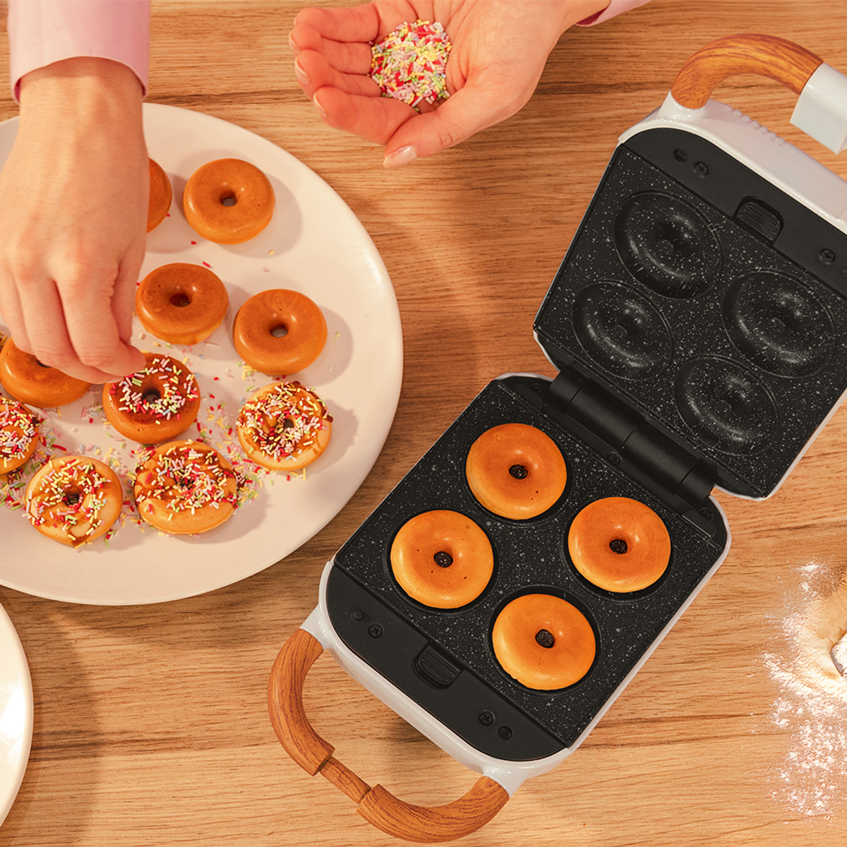 Una persona decora rosquillas con chispas. Las rosquillas están en un plato y en una máquina para hacer rosquillas. La escena es en una mesa de madera.