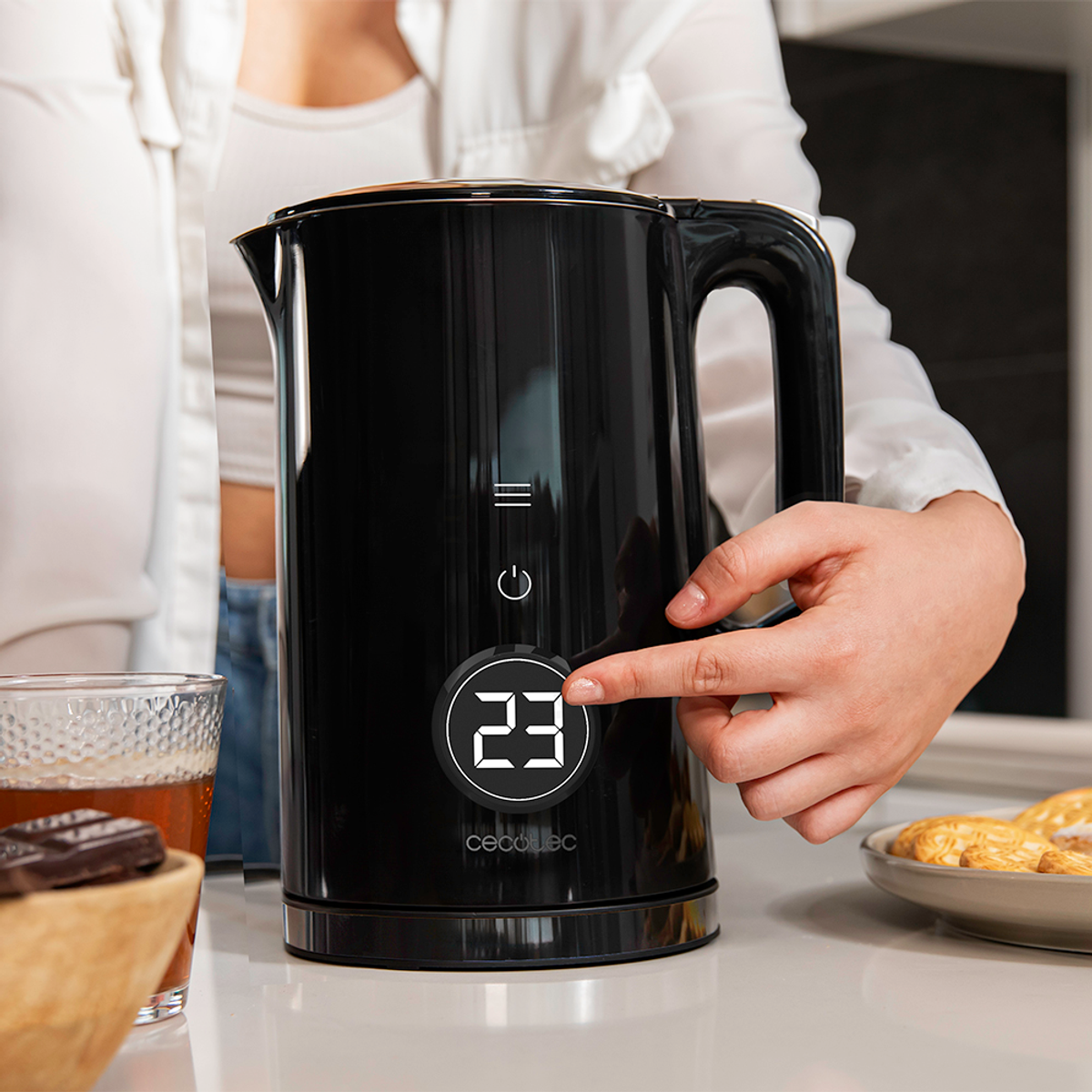 Eine Person benutzt einen schwarzen Wasserkocher. Das Display zeigt '23'. Ein Glas Tee, Schokolade und Kekse sind sichtbar.