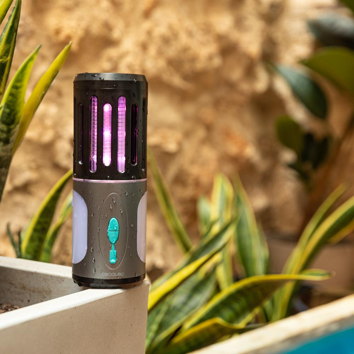Ein schwarz-grauer Insektenvernichter. Es hat ein violettes Licht im Inneren. Umgeben von grünen Pflanzen und einer beigen Steinmauer.