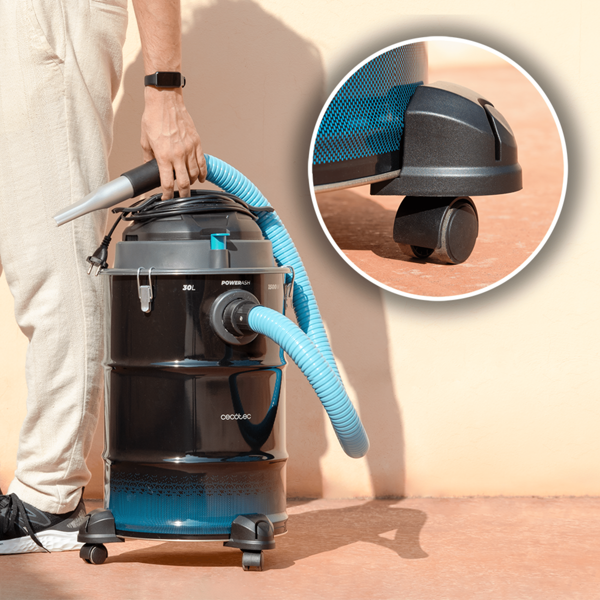 Una persona tiene in mano un aspirapolvere nero e blu con un tubo blu. Lo sfondo è un muro beige e un pavimento marrone.