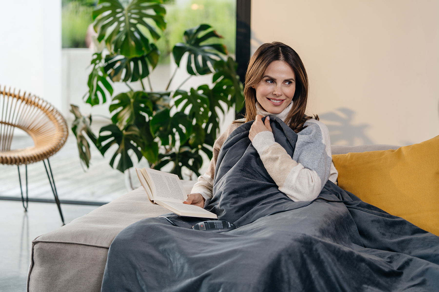 Eine Frau sitzt auf einem Sofa und liest ein Buch. Sie ist mit einer dunklen Decke bedeckt, lächelt und hat eine Pflanze im Hintergrund.