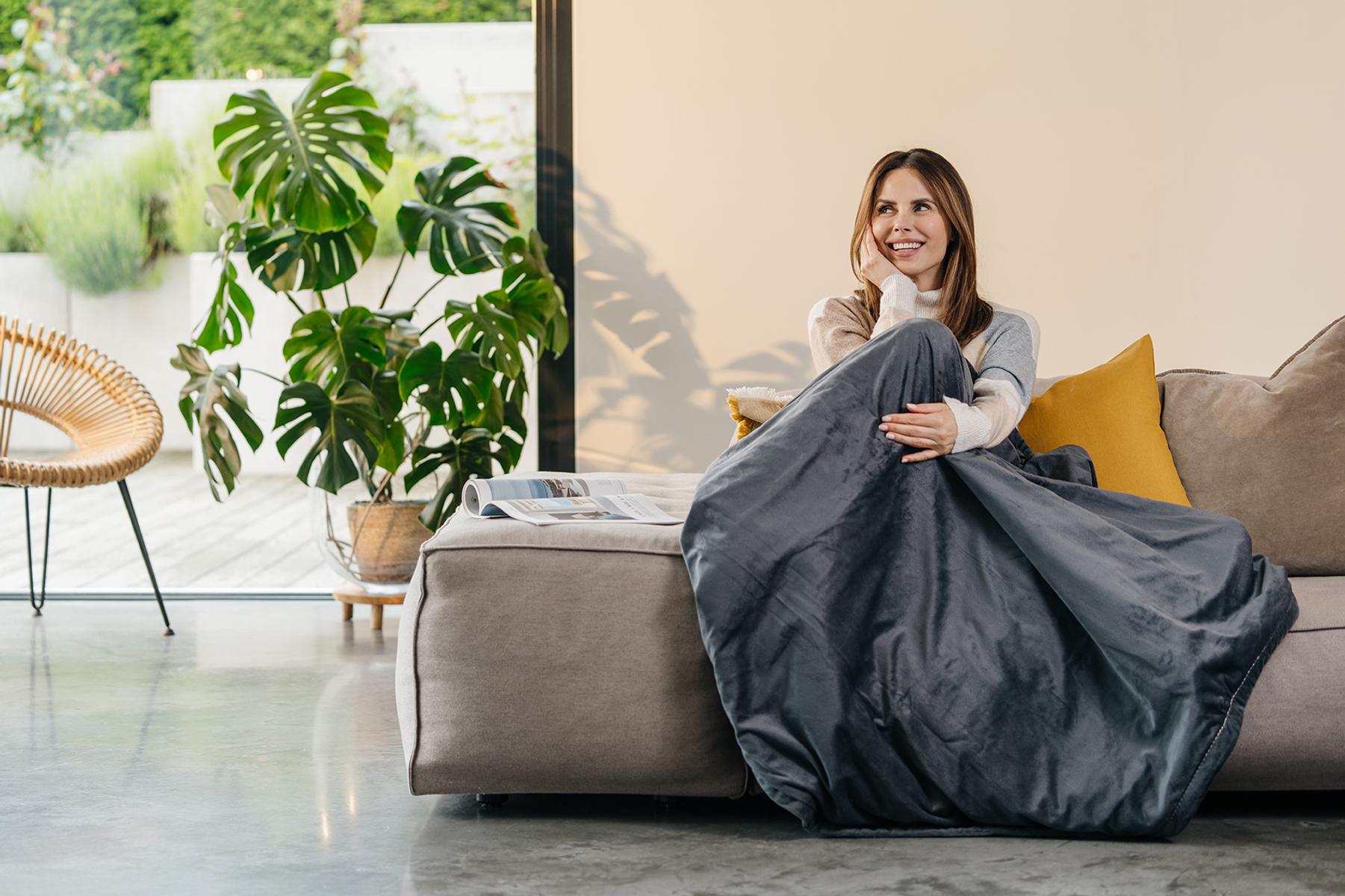 Frau lächelt, in eine graue Decke gehüllt, auf einem Sofa. Eine große Pflanze und ein Stuhl sind in der Nähe. Der Hintergrund ist ein helles Fenster.