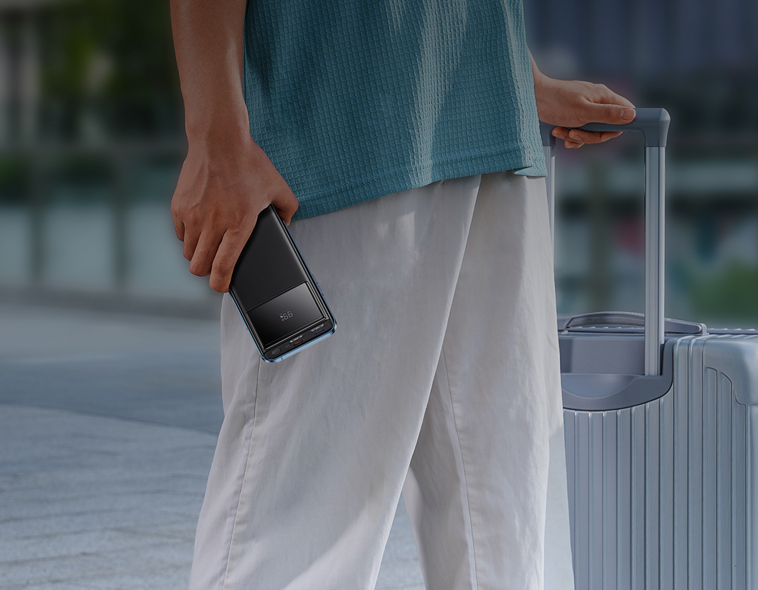 Persona che tiene in mano un telefono nero, che tira una valigia. Indossa una camicia verde acqua e pantaloni bianchi, cammina sul marciapiede.