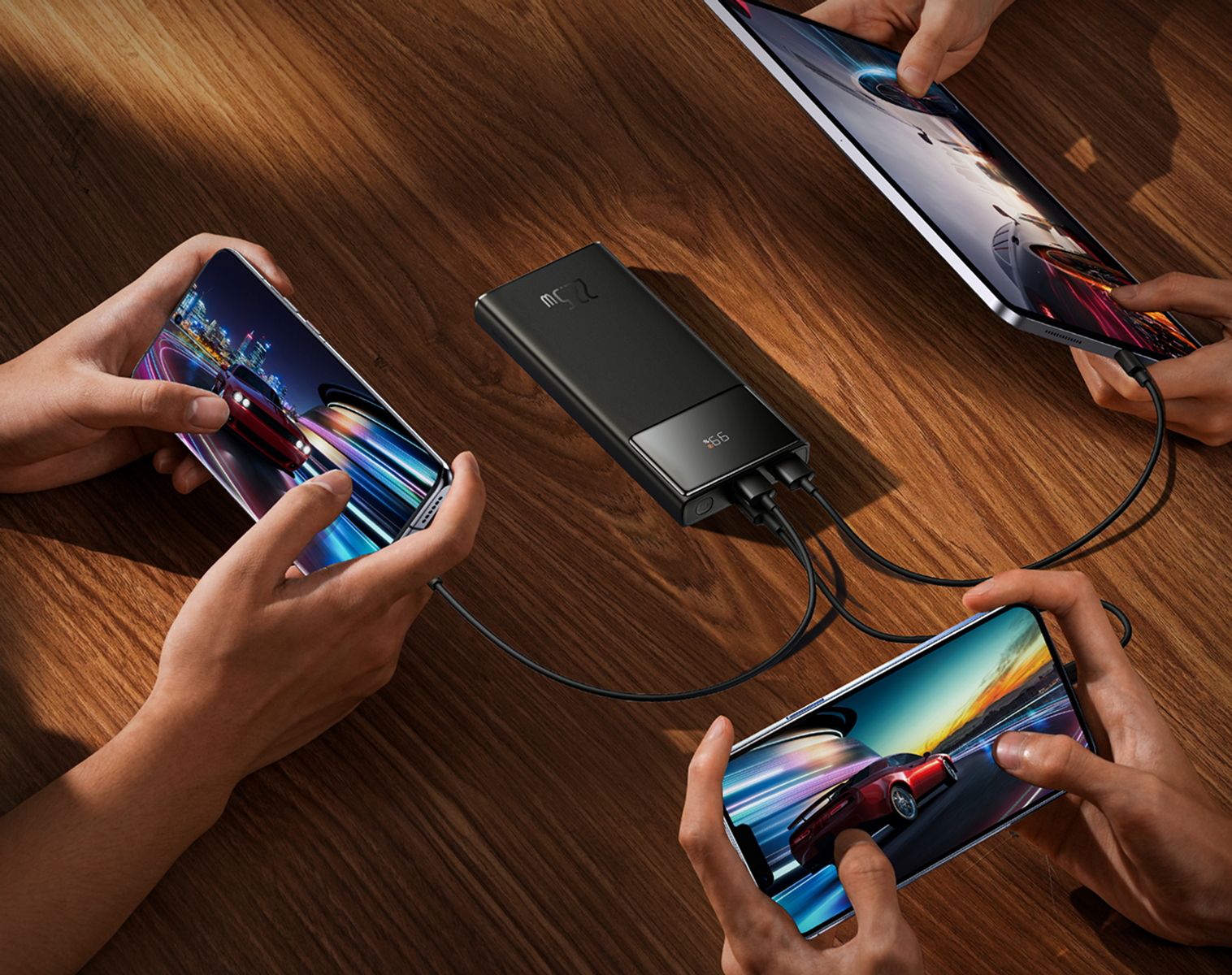 Le mani tengono telefoni e un tablet, tutti collegati a un power bank nero su un tavolo di legno. Gli schermi mostrano giochi di corse.
