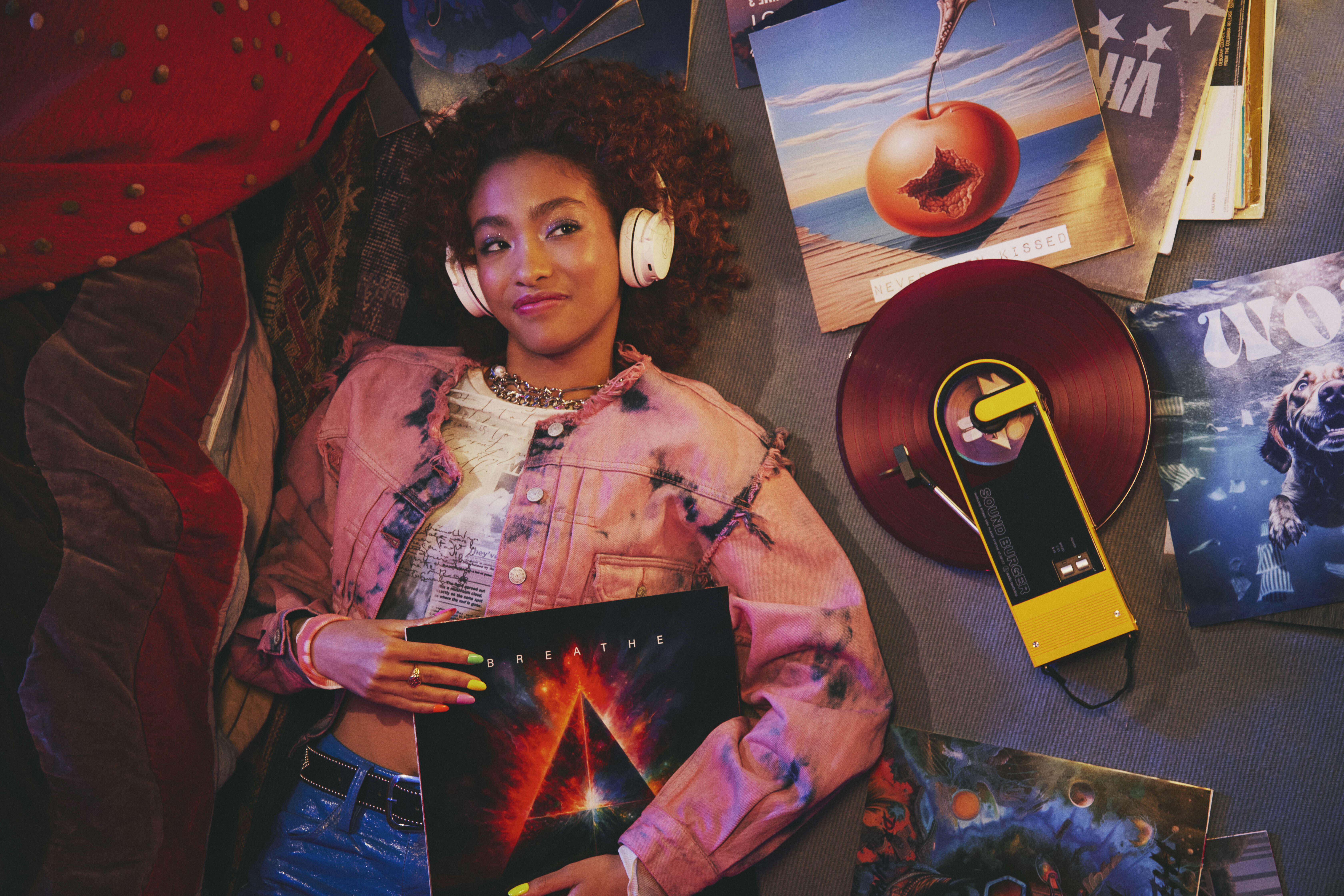 Eine Frau mit Kopfhörern und einer rosa Jacke ist von Schallplatten umgeben. Sie hält ein Album mit dem Titel 'Breathe'.