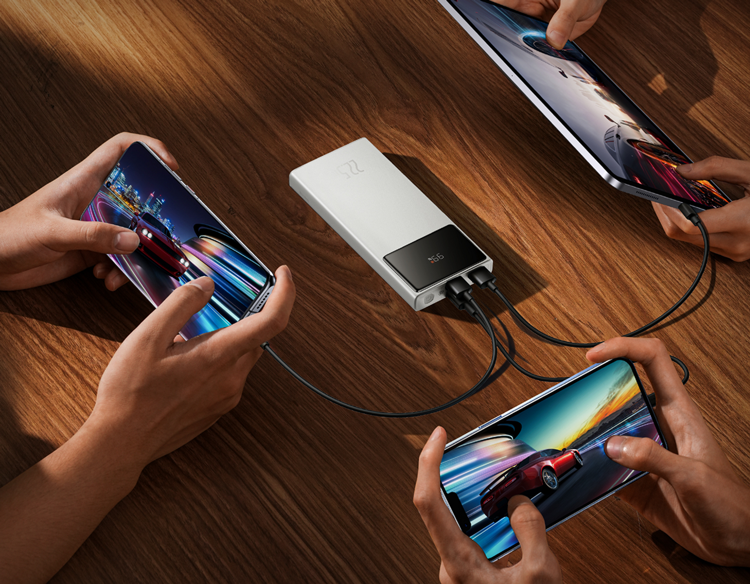 Le mani tengono smartphone e tablet, con giochi di corse. Un power bank carica i dispositivi su un tavolo di legno.