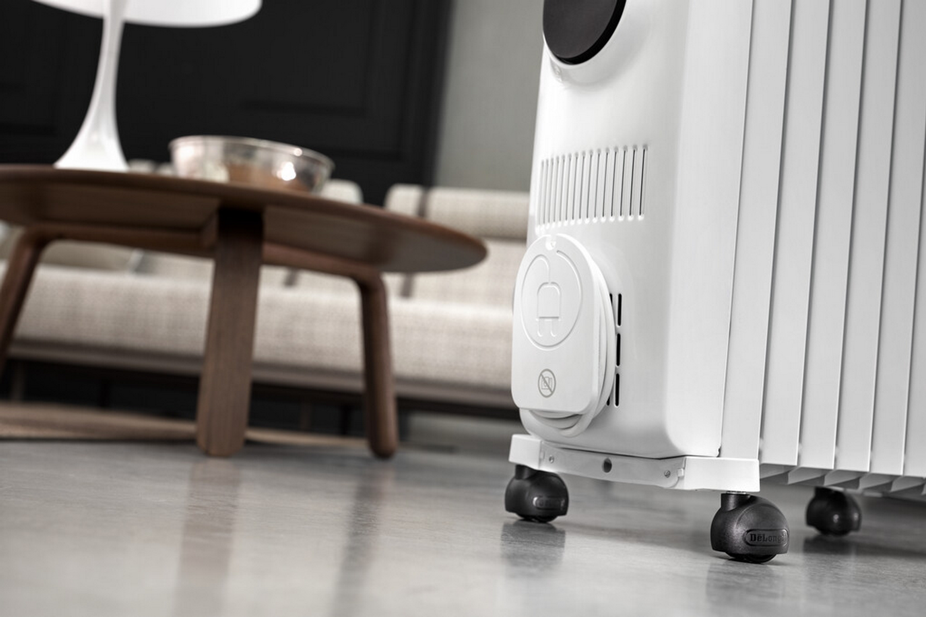 Ein weißer Ölradiator auf Rädern. Ein Tisch und ein Sofa sind im Hintergrund. Der Boden ist glänzend.