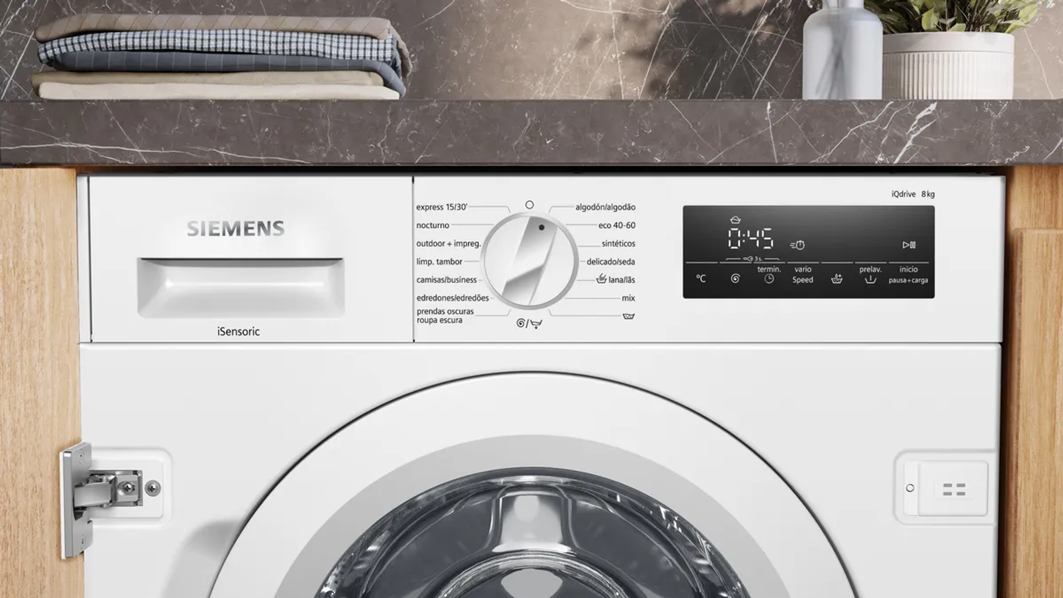 Una lavadora Siemens blanca con panel de control, dial y pantalla digital. La ropa está apilada encima.