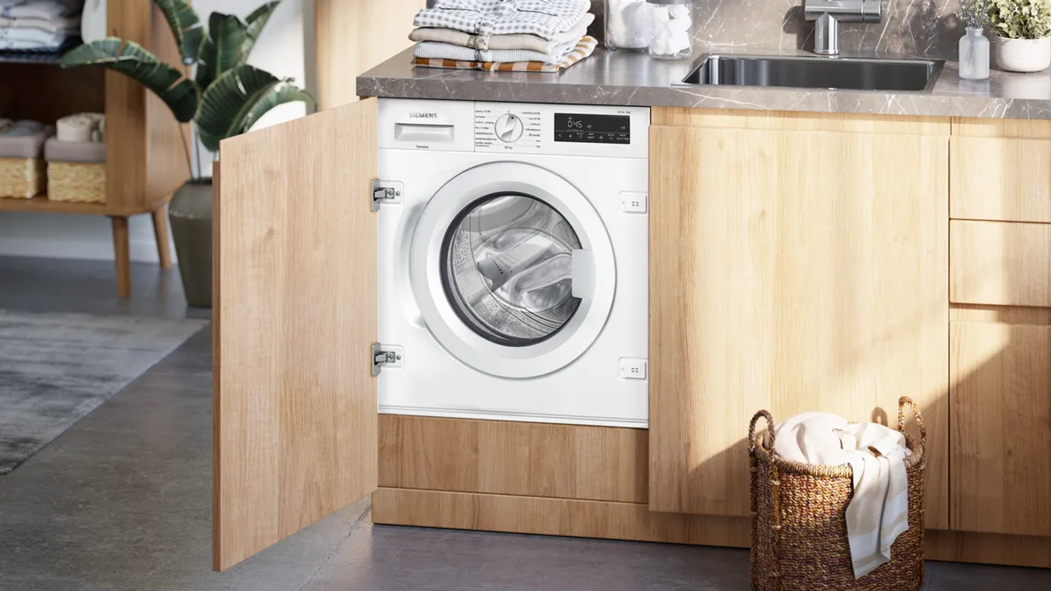 Una lavadora Siemens blanca está en un armario de madera. Se ve una cesta con ropa y un fregadero.