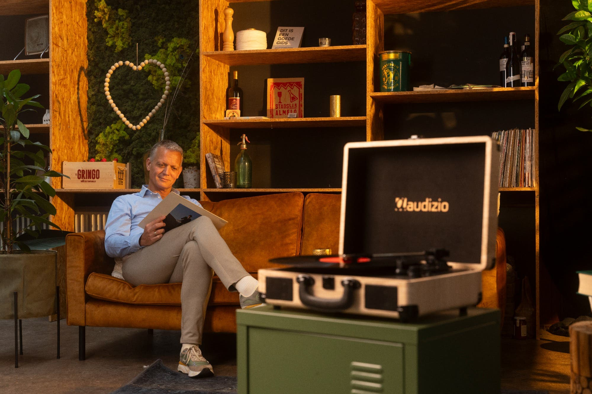 Hombre sentado en sofá leyendo. Tocadiscos en gabinete verde. Estanterías con objetos. Decoración de corazón de madera.