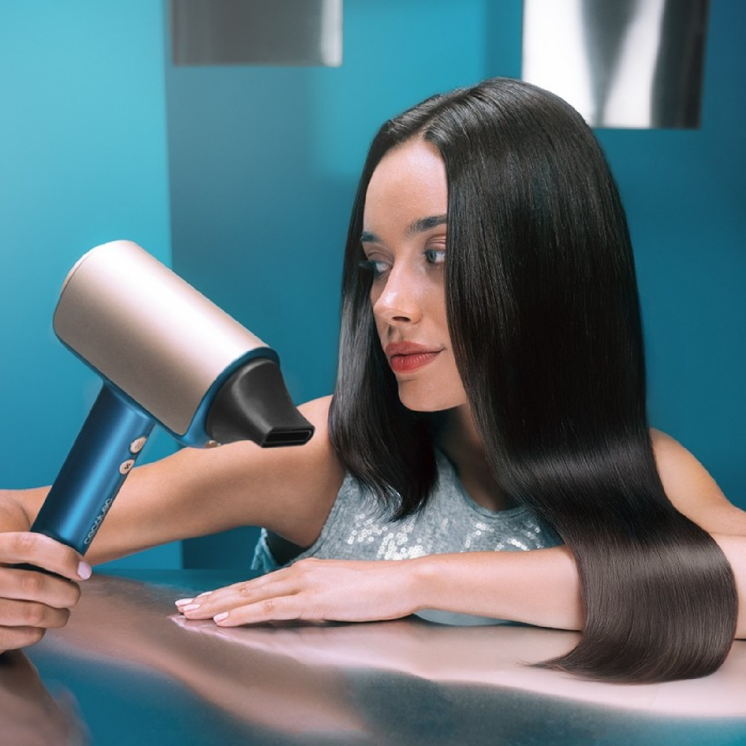 Een vrouw kijkt naar een föhn, haar lange zwarte haar stroomt. De föhn is blauw en goud. De achtergrond is blauwgroen.