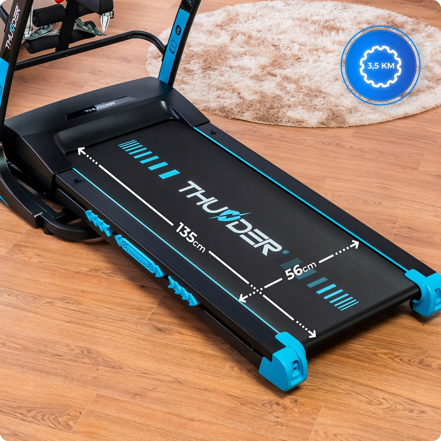 Czarna bieżnia z niebieskimi akcentami na drewnianej podłodze. Dywan w tle. Bieżnia ma napis 'THUNDER'.