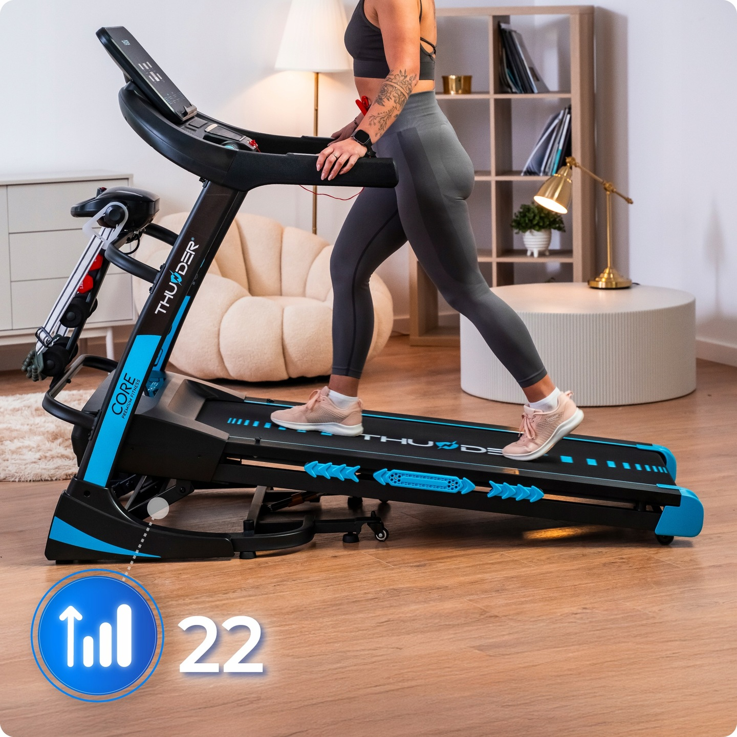 Kobieta ćwiczy na bieżni. Bieżnia jest czarno-niebieska. Kobieta ma na sobie strój do ćwiczeń, a w pokoju są meble.