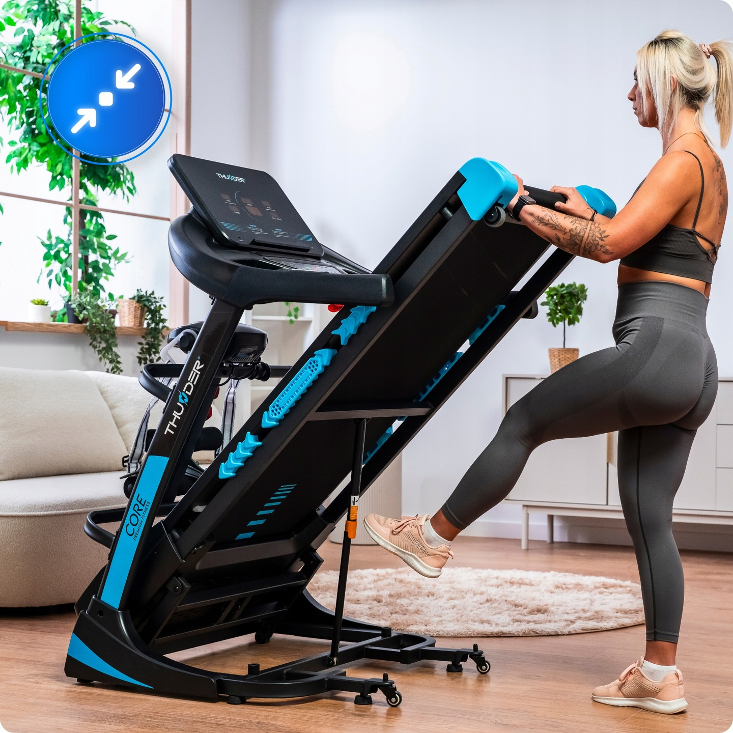 Kobieta ćwiczy na bieżni. Maszyna jest czarno-niebieska. Jest w pokoju z oknem i białą sofą.