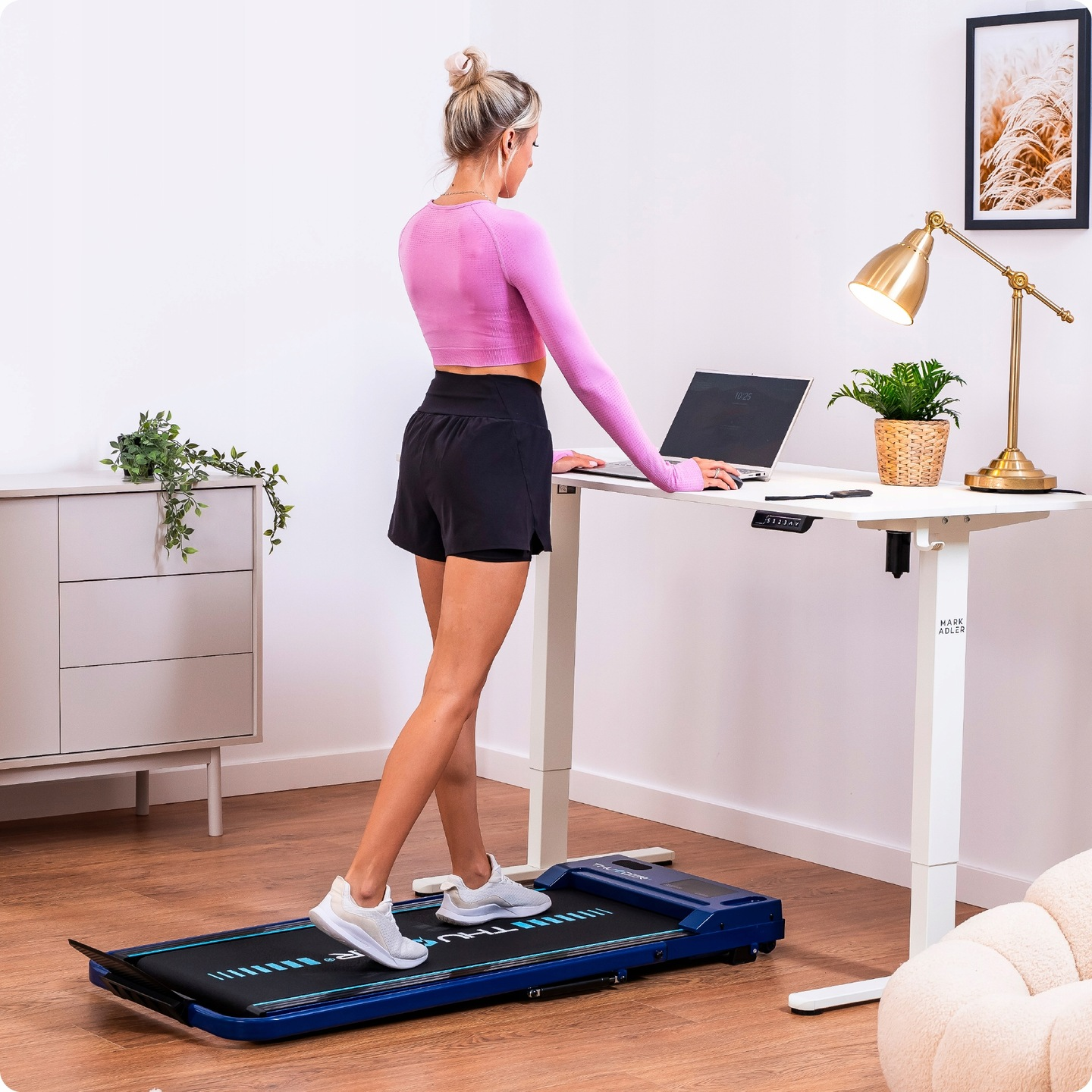 Kobieta w różowym topie chodzi po biurku z bieżnią. Laptop, lampa i roślina na biurku. Szafka i grafika w tle.