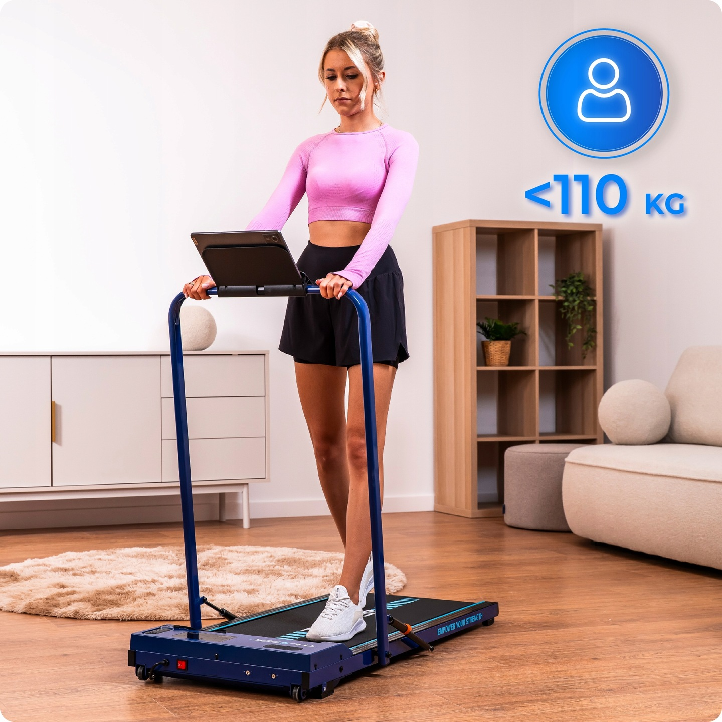 Kobieta ćwiczy na bieżni. Ma na sobie różowy top i czarne szorty. Tło z meblami i znakiem ograniczenia wagi.