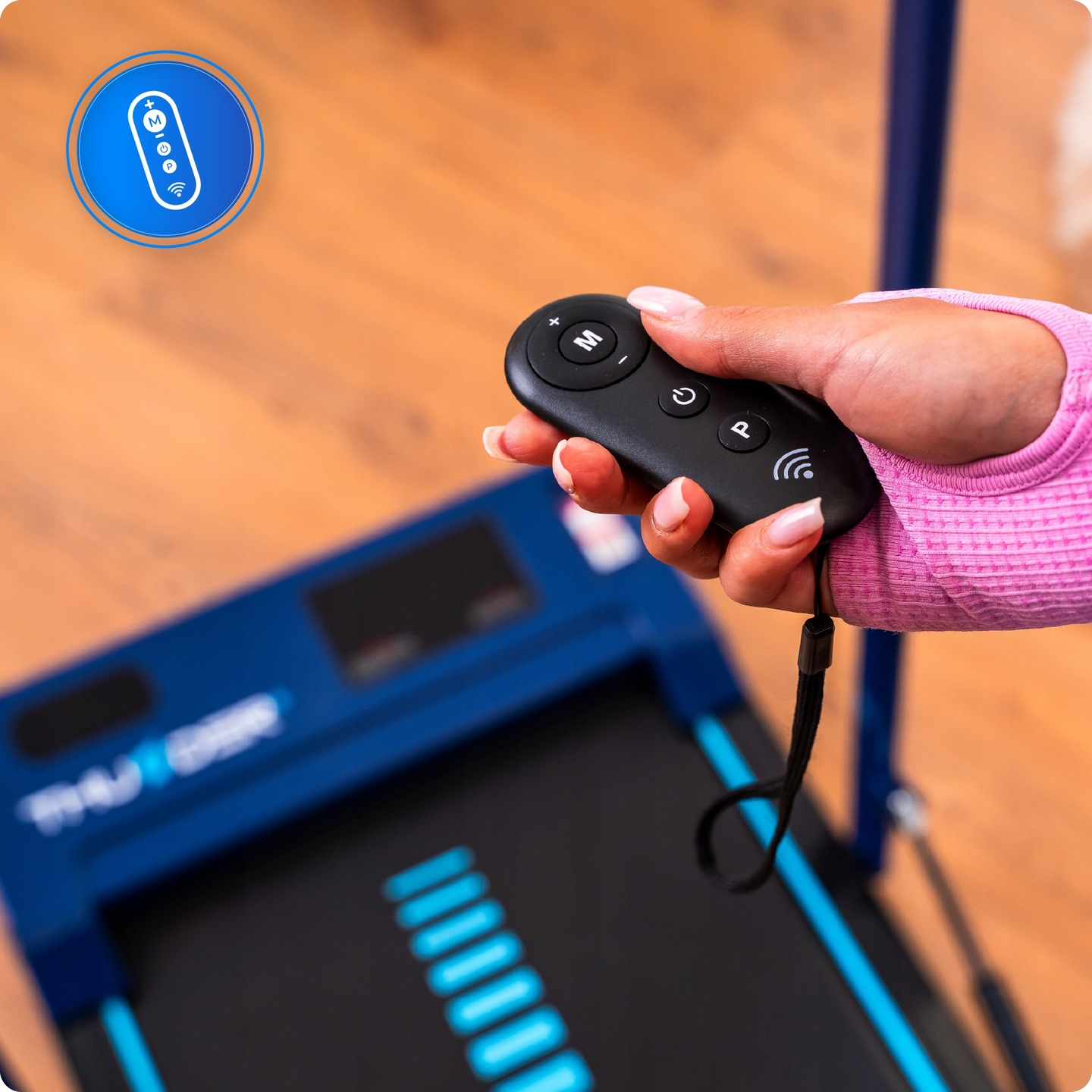 Dłoń trzyma czarnego pilota z przyciskami. Bieżnia jest niebiesko-czarna. Niebieskie koło pokazuje ikony pilota.