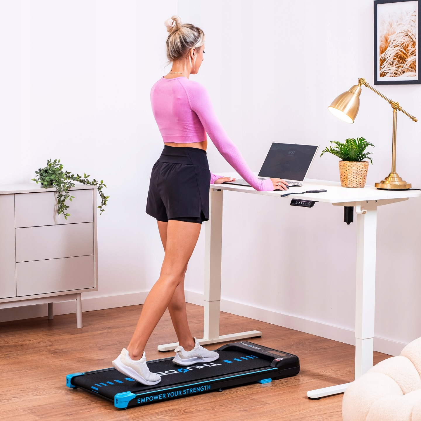 Kobieta w stroju do ćwiczeń korzysta z biurka z bieżnią. Otwarty laptop, złota lampa, roślina i grafika. Drewniana podłoga. Białe biurko i ściana.