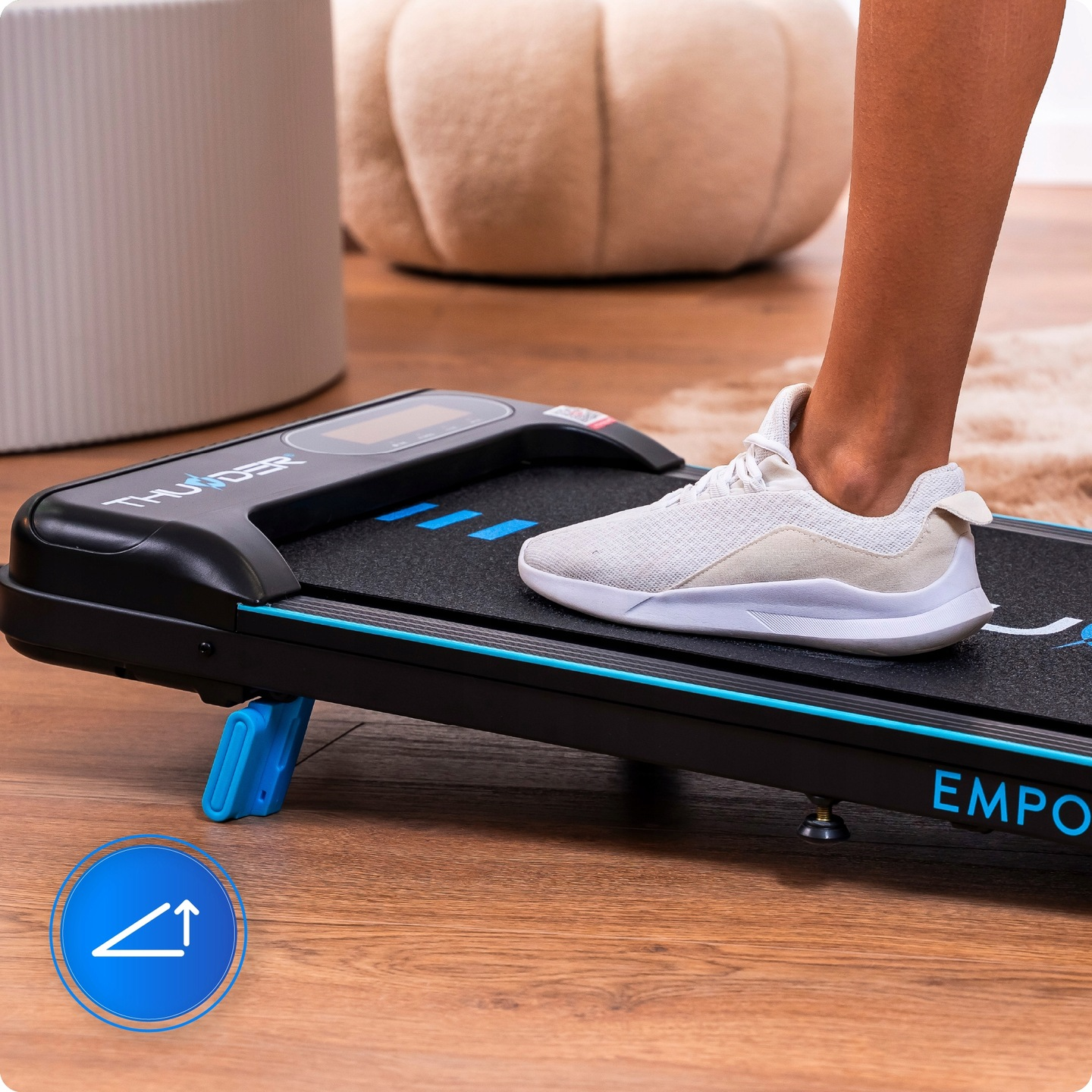 Stopa osoby w białym bucie na czarnej bieżni. Widoczne są niebieskie akcenty i niebieski przycisk. Na tle beżowa poduszka.