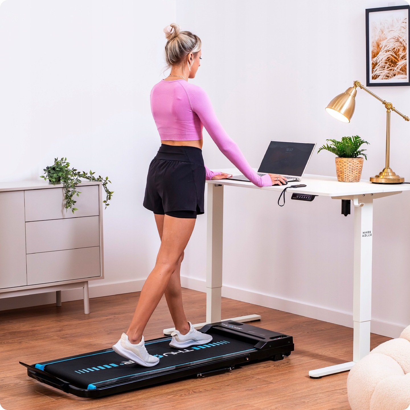 Kobieta ćwiczy na bieżni podczas pracy przy biurku z laptopem. Na biurku stoi lampa i roślina.