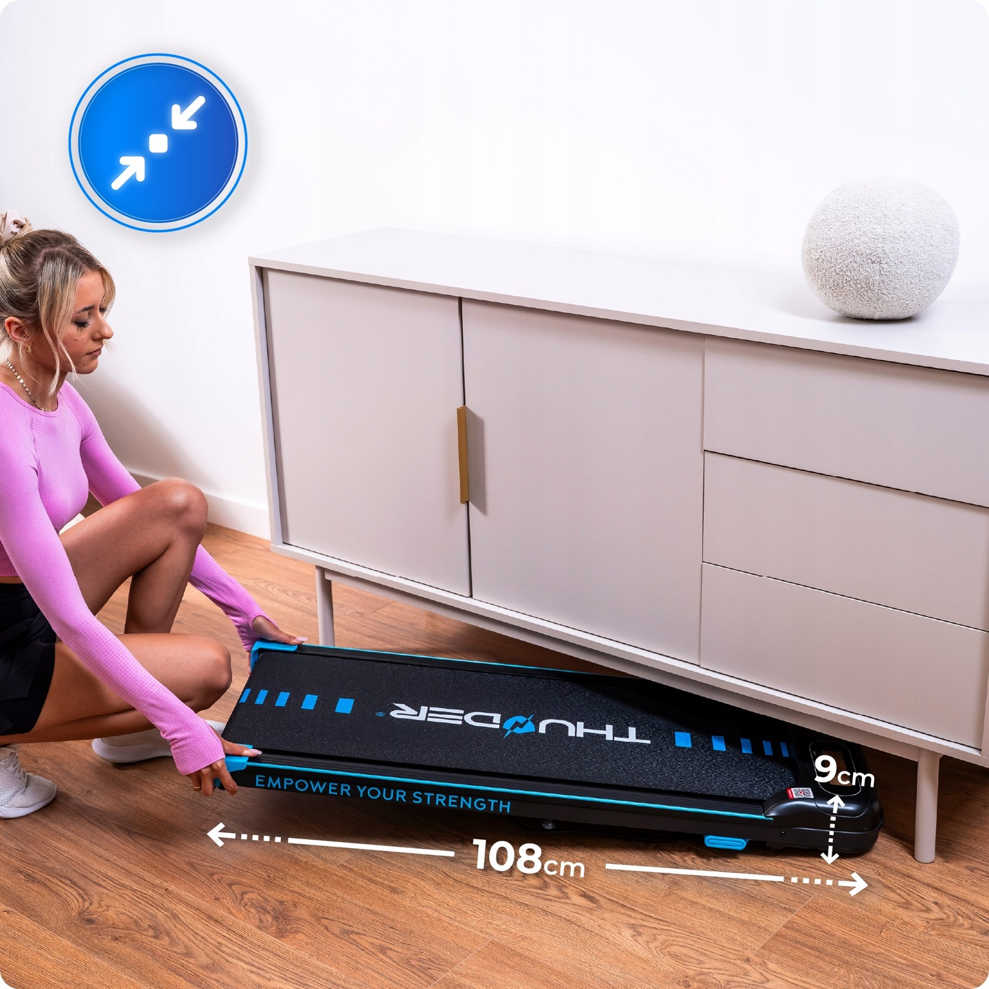 Kobieta umieszcza czarno-niebieskie urządzenie do ćwiczeń pod szafką. Urządzenie ma 108 cm długości i 9 cm wysokości.