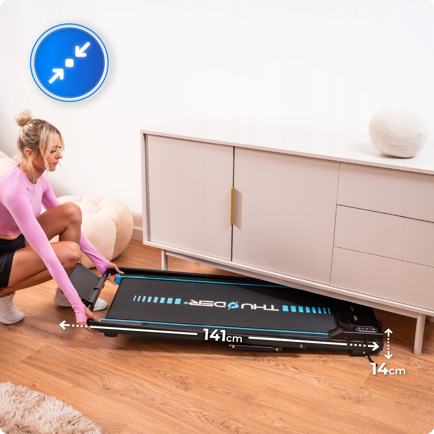 Kobieta wyciąga czarną bieżnię spod białej szafki. Bieżnia ma niebieskie akcenty, a wymiary są wyświetlane.