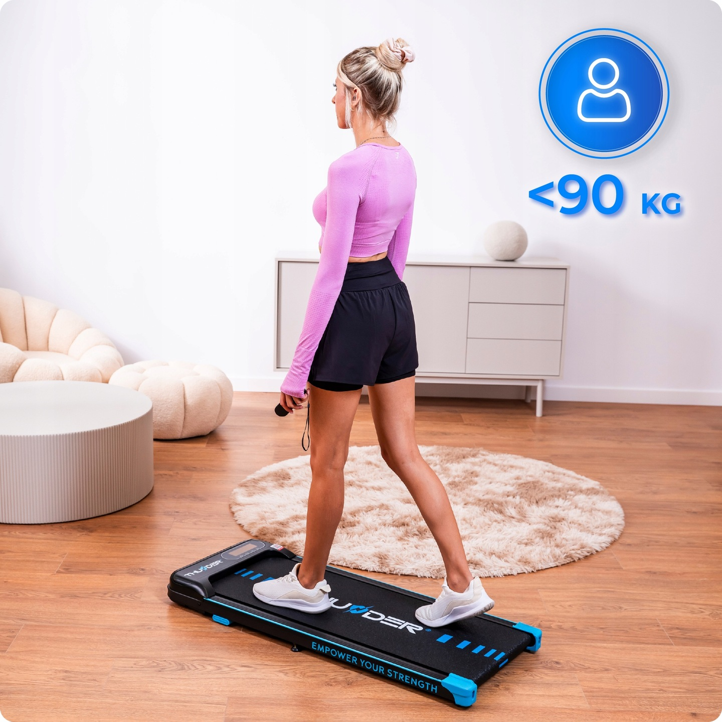 Kobieta idzie na bieżni w pomieszczeniu. Ma na sobie różowy top i czarne szorty. Widoczny jest znak ograniczenia wagi.