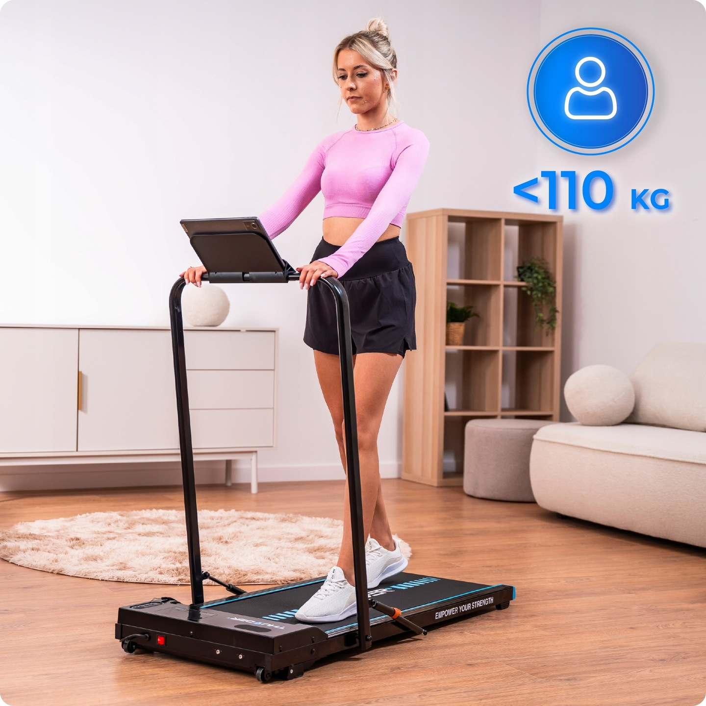 Kobieta ćwiczy na bieżni. Ma na sobie różowy top i czarne szorty. Znak wskazuje limit wagowy poniżej 110 kg.