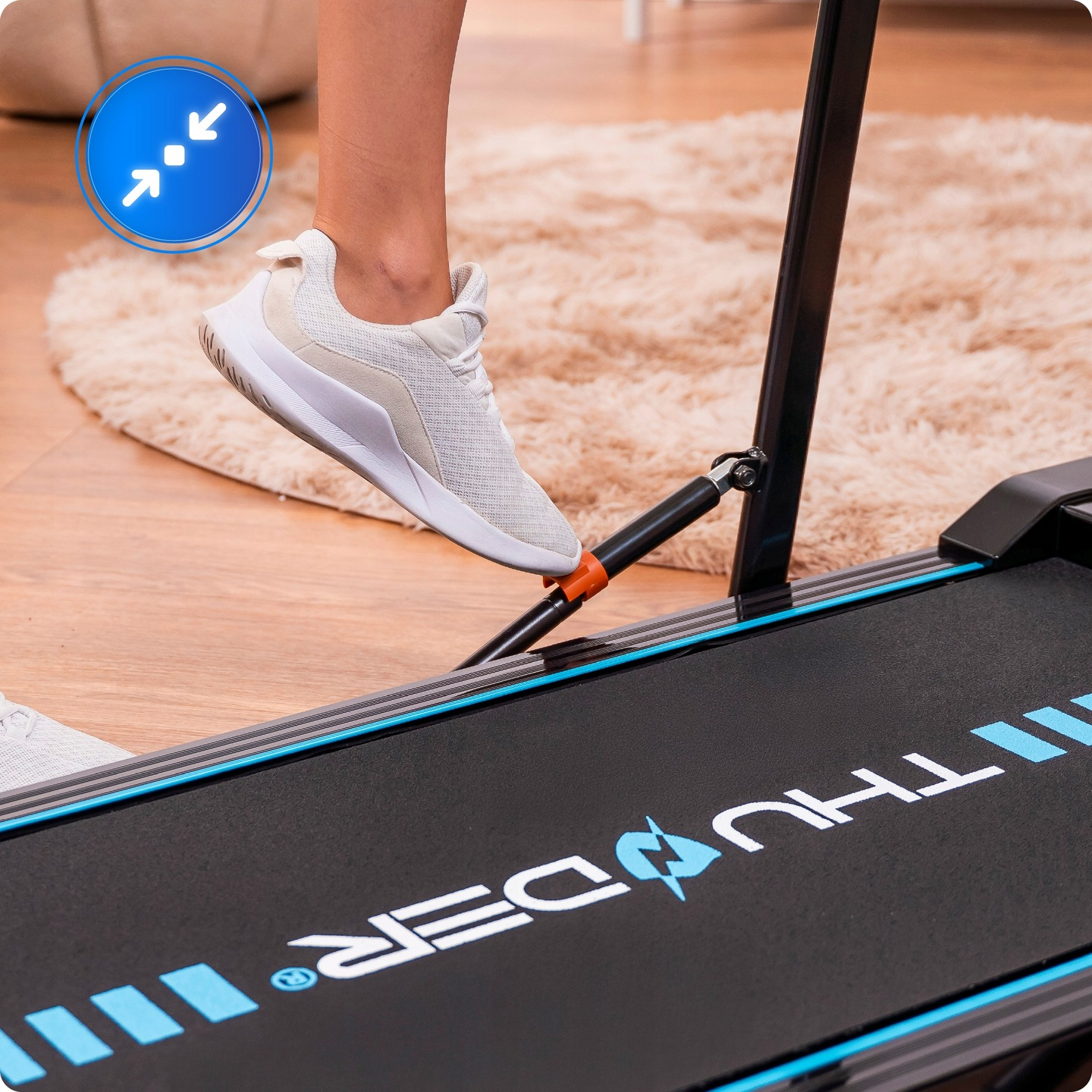 Stopa osoby w białym bucie stąpa na bieżnię. Bieżnia jest czarna z niebieskimi akcentami i nazwą marki. W tle puszysty dywan.