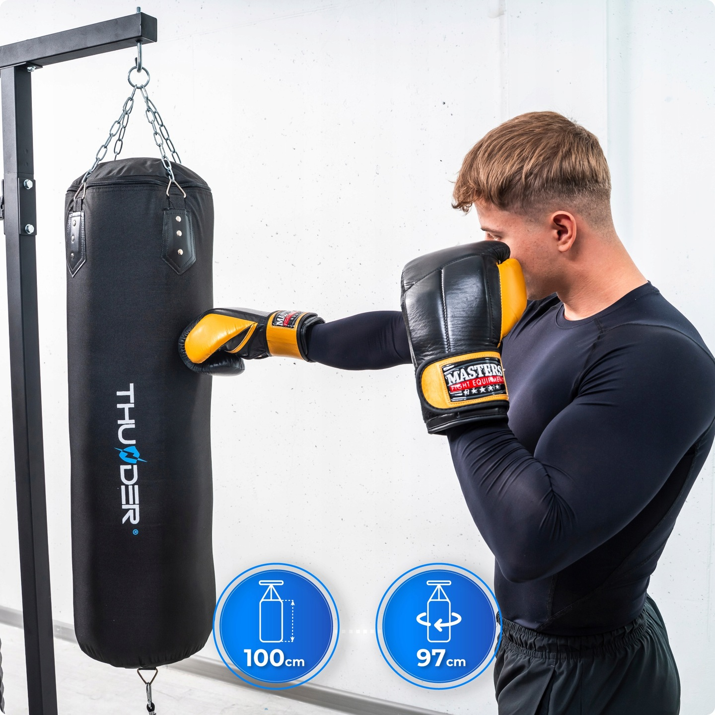 Mężczyzna w czarnym stroju sportowym uderza w czarny worek bokserski żółto-czarnymi rękawicami bokserskimi. Worek wisi na czarnej ramie.