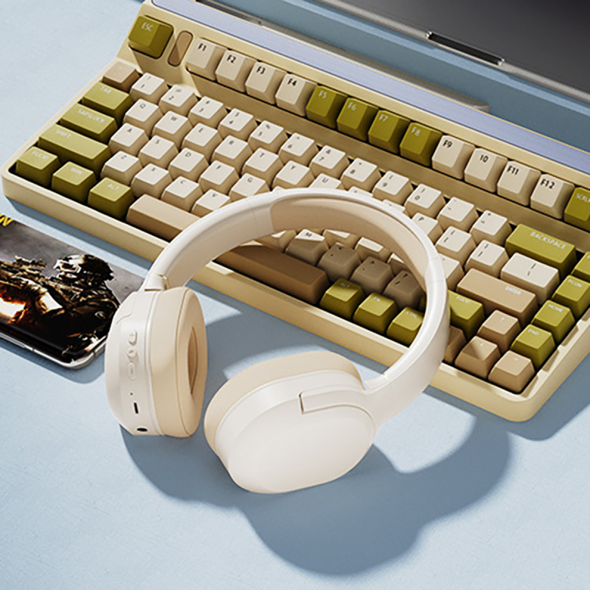 Ein cremefarbener drahtloser Kopfhörer liegt auf einer blauen Oberfläche in der Nähe einer Tastatur mit cremefarbenen und grünen Tasten und einem Smartphone.