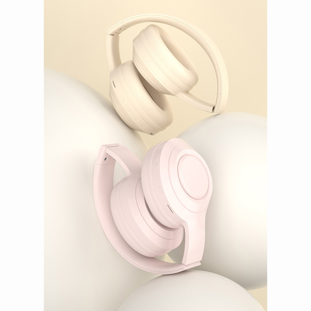 Zwei zusammengeklappte Kopfhörer, einer rosa und einer beige, liegen auf weißen, abgerundeten Objekten. Der Hintergrund ist hellbeige.