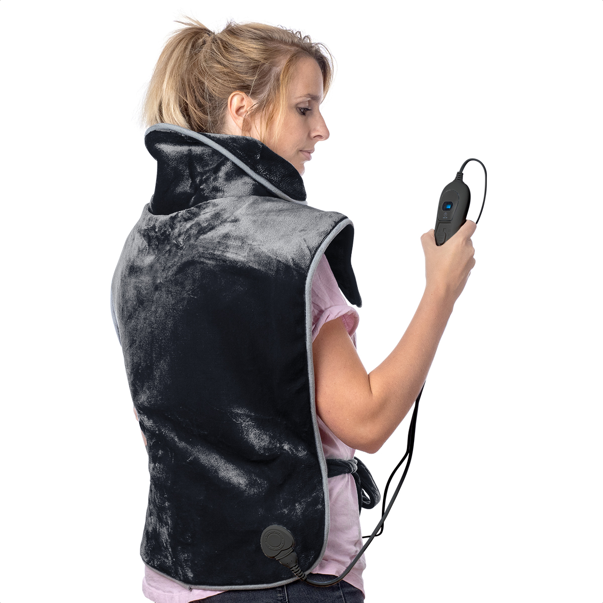 Woman wearing a black heating pad, holding a remote. The pad is on her shoulders and back. She is looking at the remote.
