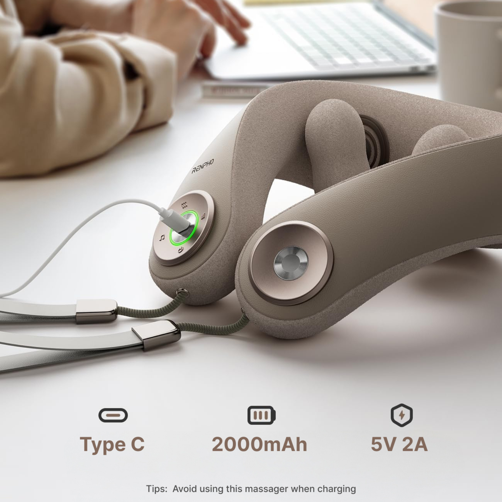 Een beige nekmassager is aangesloten op een wit oppervlak. De massager heeft knoppen en luidsprekers. Op de achtergrond werkt een persoon op een laptop.