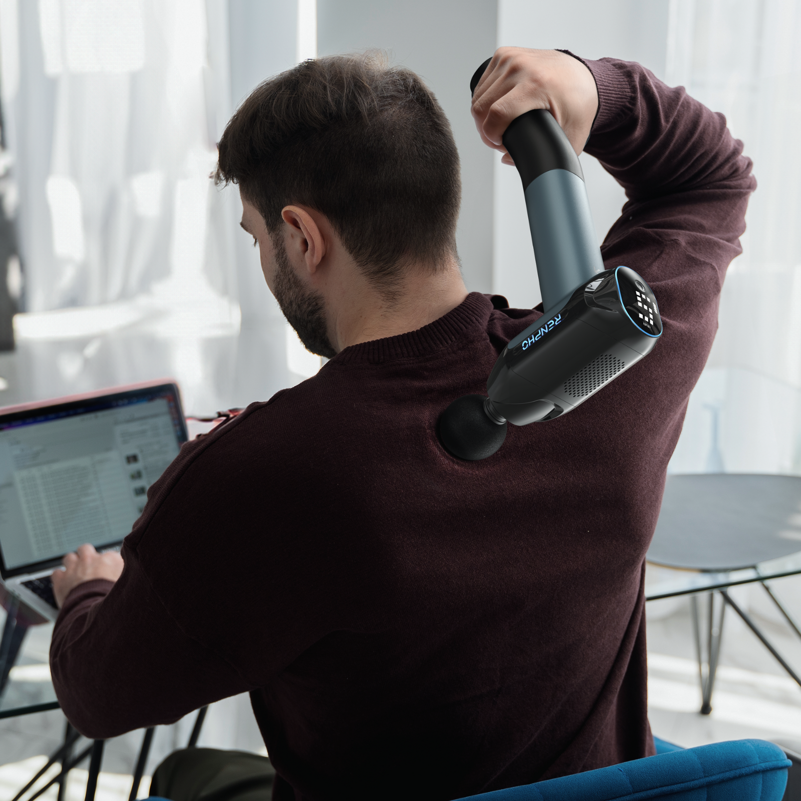 Mann benutzt Massagepistole am Rücken, während er am Laptop arbeitet. Burgunderfarbener Pullover, blauer Stuhl, Fenster im Hintergrund.