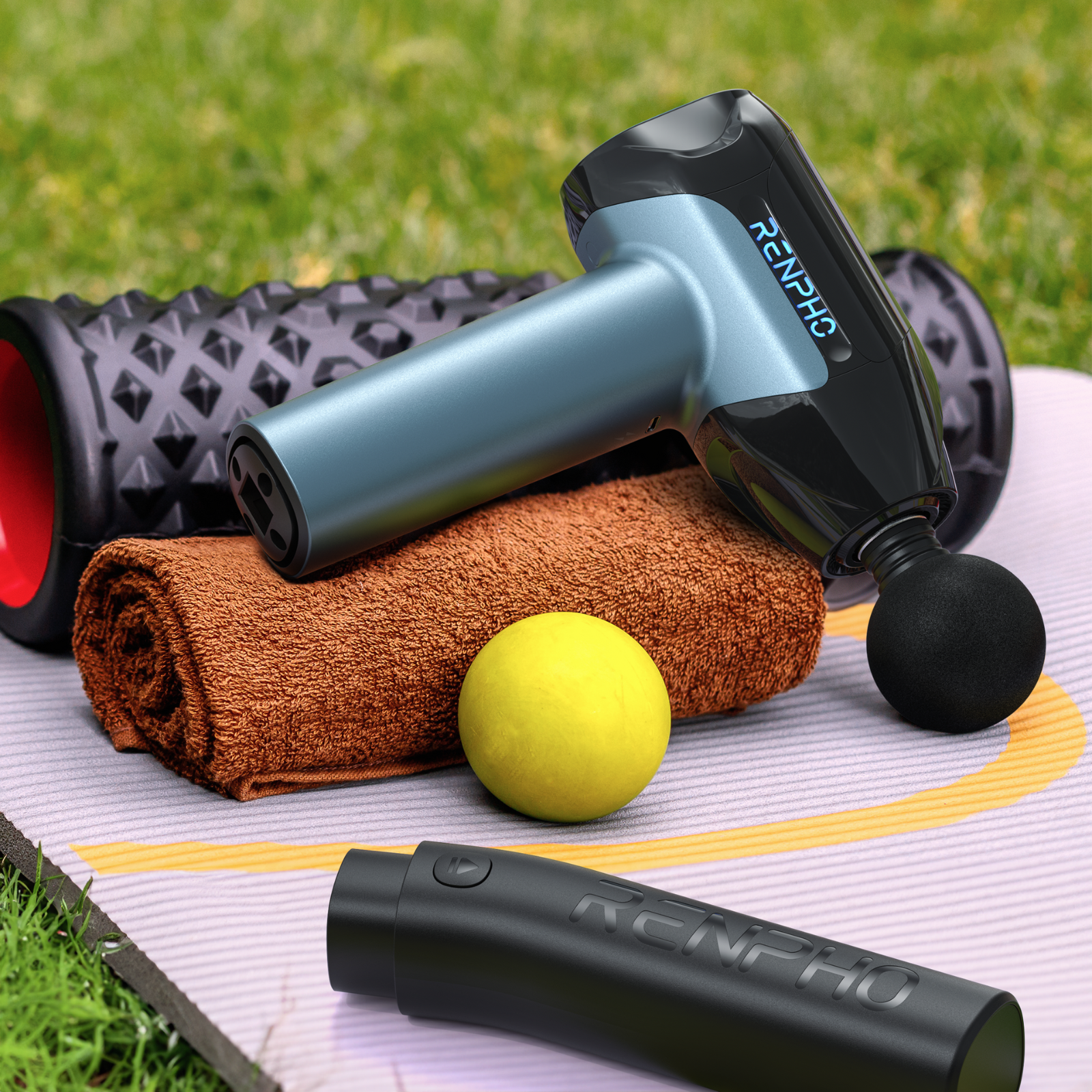 Massagepistole, Rolle, Handtuch, gelber Ball und ein weiteres Gerät auf einer Matte, alles auf Gras.