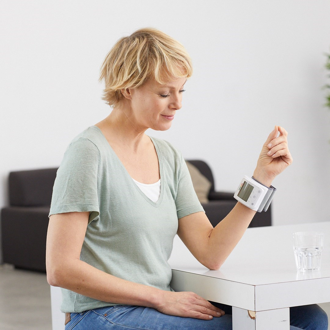 Kobieta z ciśnieniomierzem na nadgarstku, siedząca przy białym stole. Obecna jest szklanka wody.