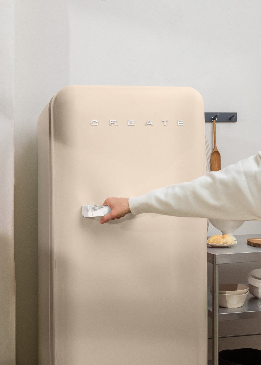Un refrigerador beige tiene la palabra CREATE. Una mano abre el refrigerador con un tirador de mármol.