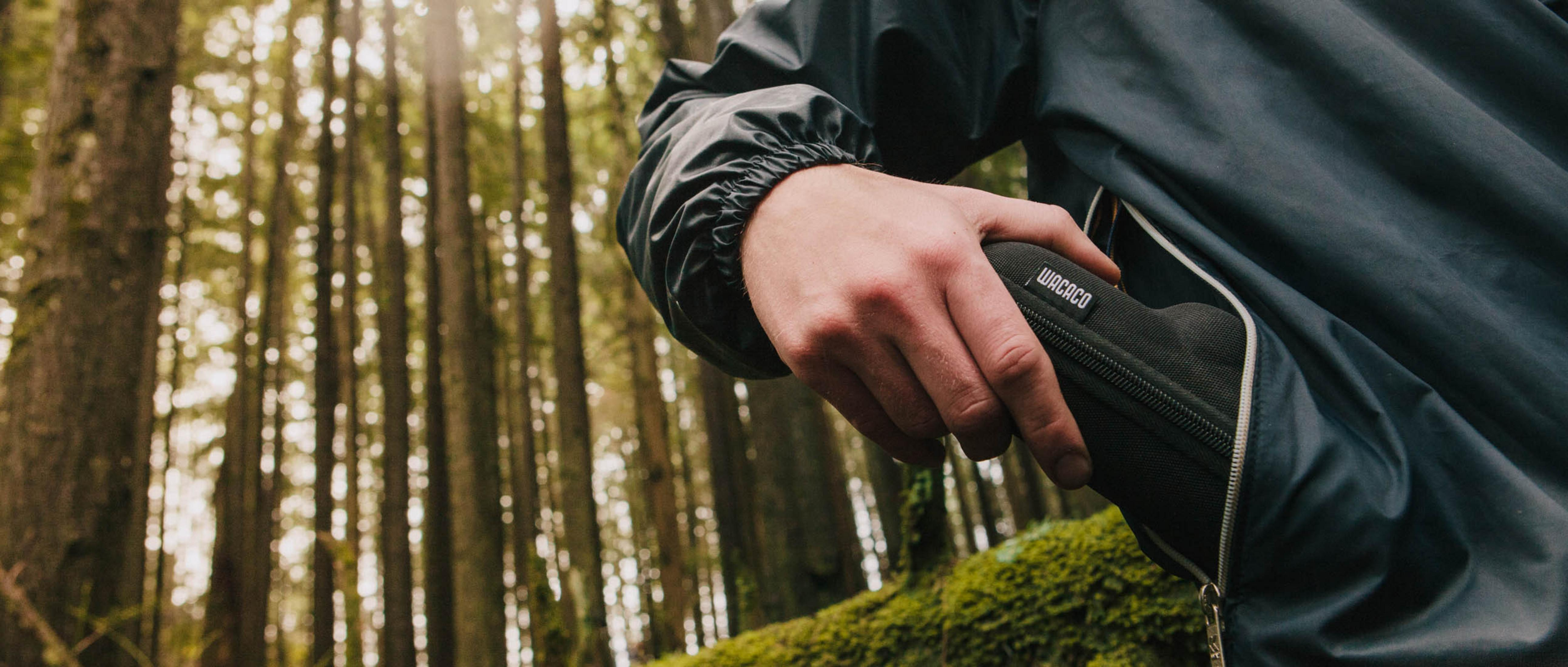 Una persona in una foresta tiene in mano una custodia nera con l'etichetta 'Wacaco'. La persona indossa una giacca scura. Gli alberi riempiono lo sfondo.