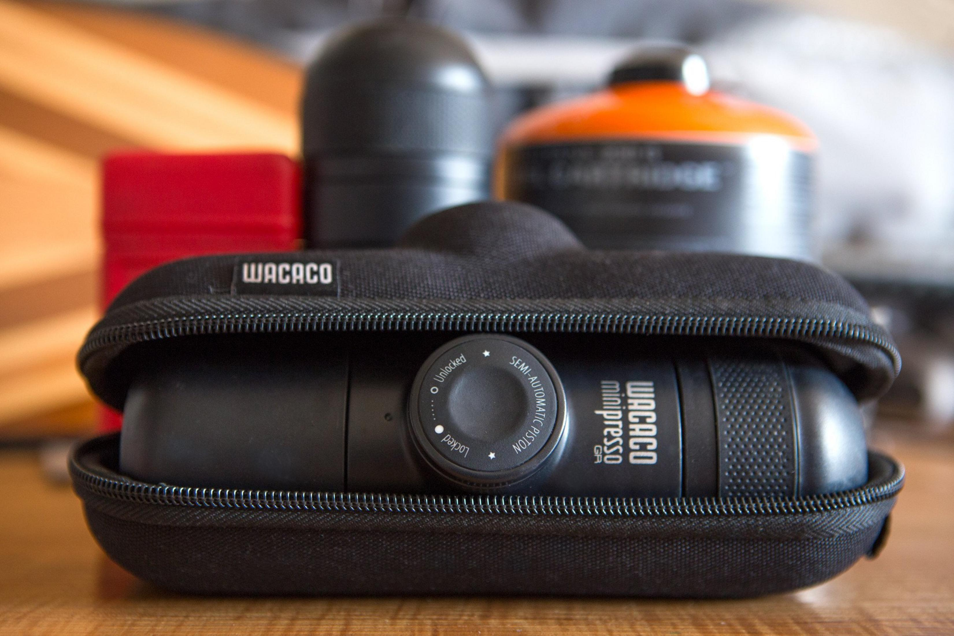 Wacaco Minipresso nero in una custodia, con altri accessori. Sullo sfondo sono visibili oggetti arancioni e rossi.