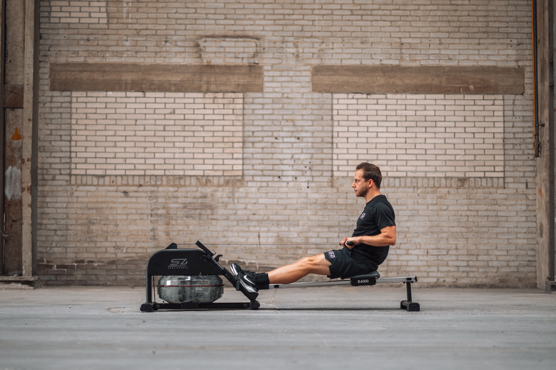 Man rowing on a machine indoors. He wears black clothes. The background is a brick wall.