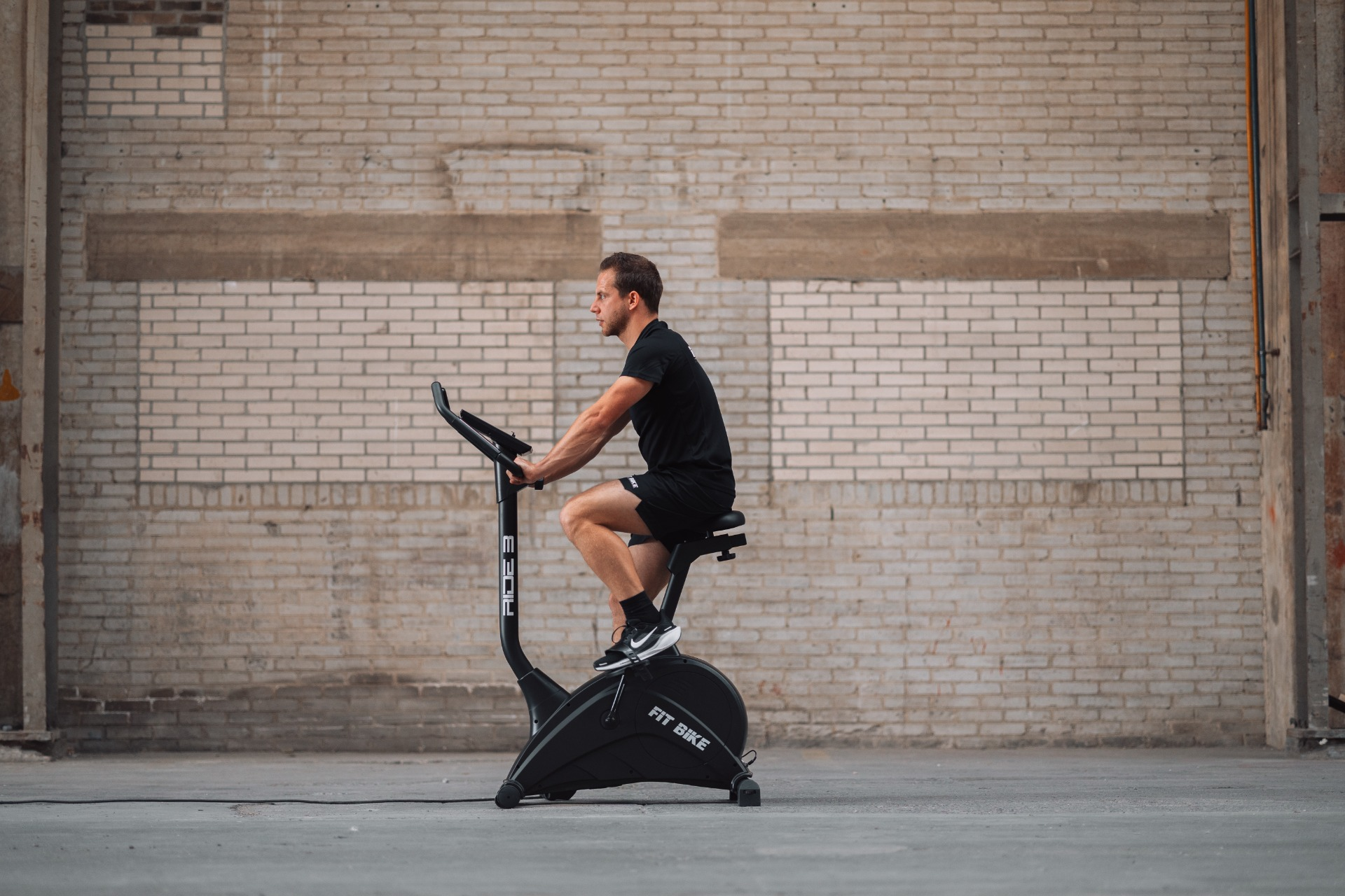 Un homme en short et chemise noirs fait du vélo sur un vélo d'appartement noir devant un mur de briques.